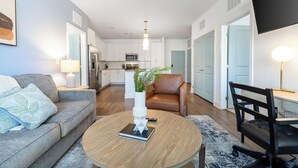 A modern living room with a grey sofa, a round coffee table, a brown leather chair, and a black office chair. There is a kitchen in the background with white cabinets and a microwave. - Landing | Beautiful 2BD, Clubhouse, Yoga Studio (Vero Beach)