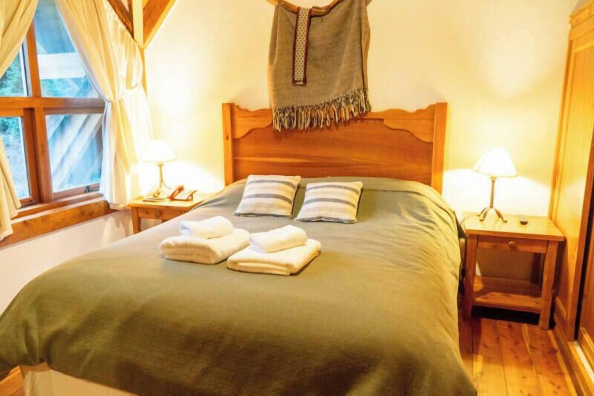 Una cama bien hecha con almohadas y toallas, cabecera de madera, lámparas de noche y una ventana con cortinas.