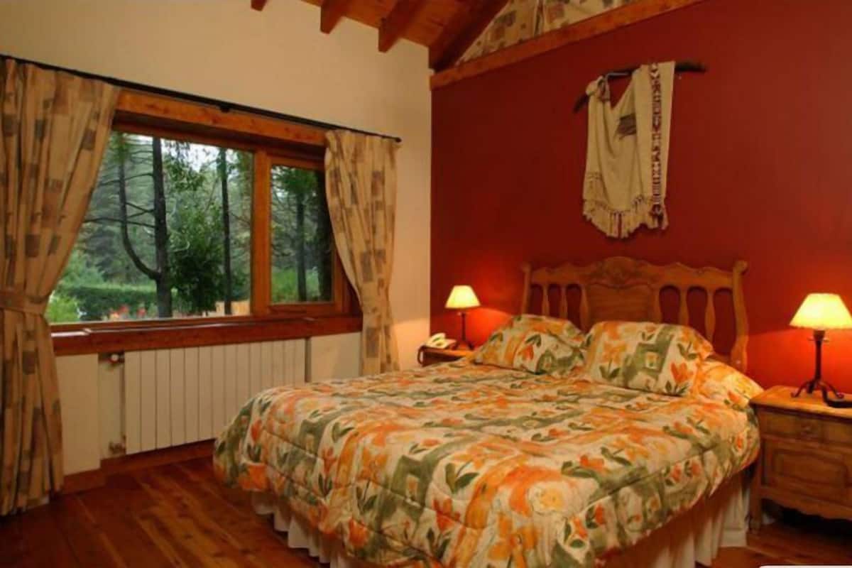 Un dormitorio con cama de madera, dos lámparas de noche, una ventana con cortinas y vistas a árboles.