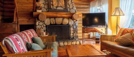 A log cabin interior with a stone fireplace, wooden furniture, and a television.