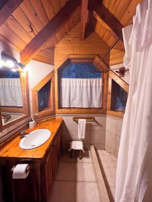 Baño de cabaña de madera con tocador de madera, lavamanos, inodoro y cortina de ducha.