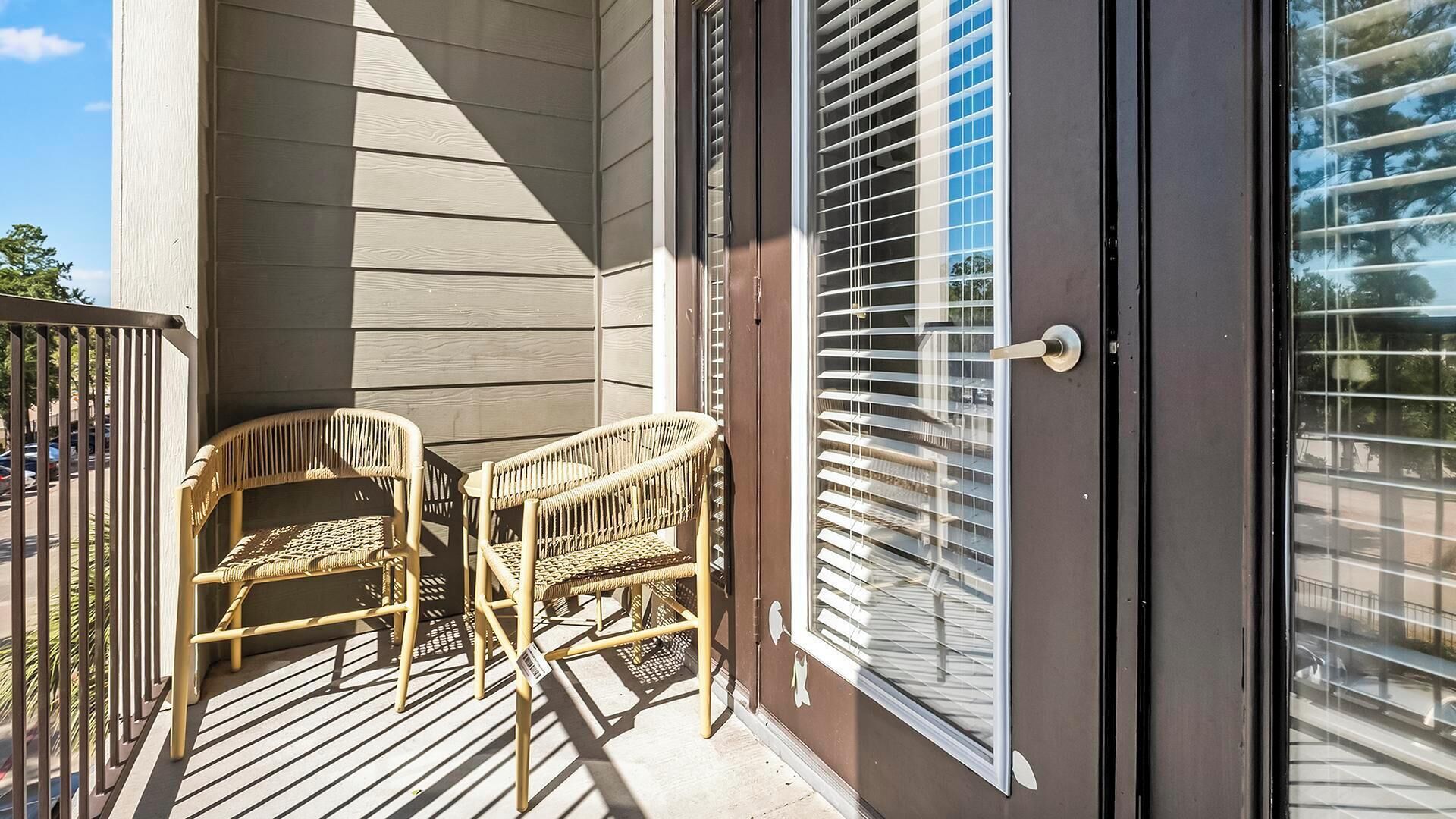 A balcony with two wicker chairs and a small table, a sliding glass door, and a view of the street.