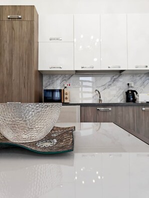 Una cucina moderna con mobili bianchi, una parete di fondo in marmo e una grande ciotola di vetro su un tavolo di vetro.