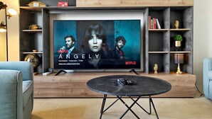 Ein Wohnzimmer mit einem großen Flachbildfernseher an einer Holzwand, einem runden Couchtisch in der Mitte und einem blauen Sessel links.
