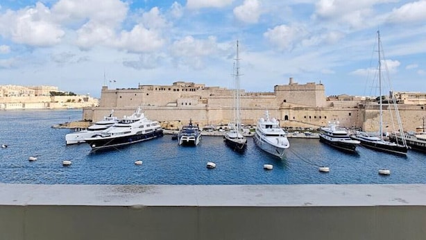 Ein Hafen mit mehreren vor Anker liegenden Yachten, ein historisches Gebäude im Hintergrund und ein strahlend blauer Himmel.
