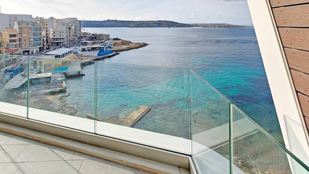 Un intérieur moderne avec un plafond en bois aux lignes courbes et un balcon en verre donnant sur une zone côtière, avec des bâtiments et une eau d’un bleu limpide.