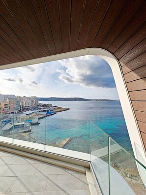 Un interno moderno con un soffitto in legno curvo e un balcone in vetro che si affaccia su una zona costiera con edifici e acque blu cristalline.