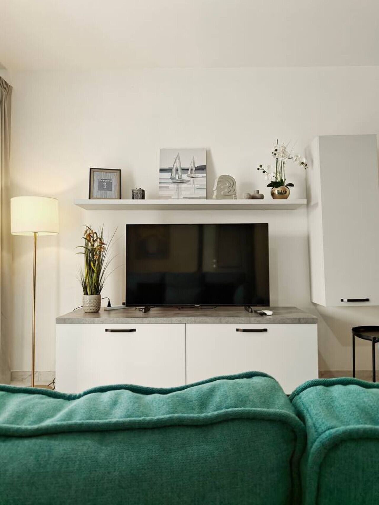 Ein modernes Hotelzimmer mit einem Flachbildfernseher auf einer weißen Kommode, einer kleinen Ablagefläche mit Dekoration, einer Topfpflanze und einer grünen Decke auf dem Bett.