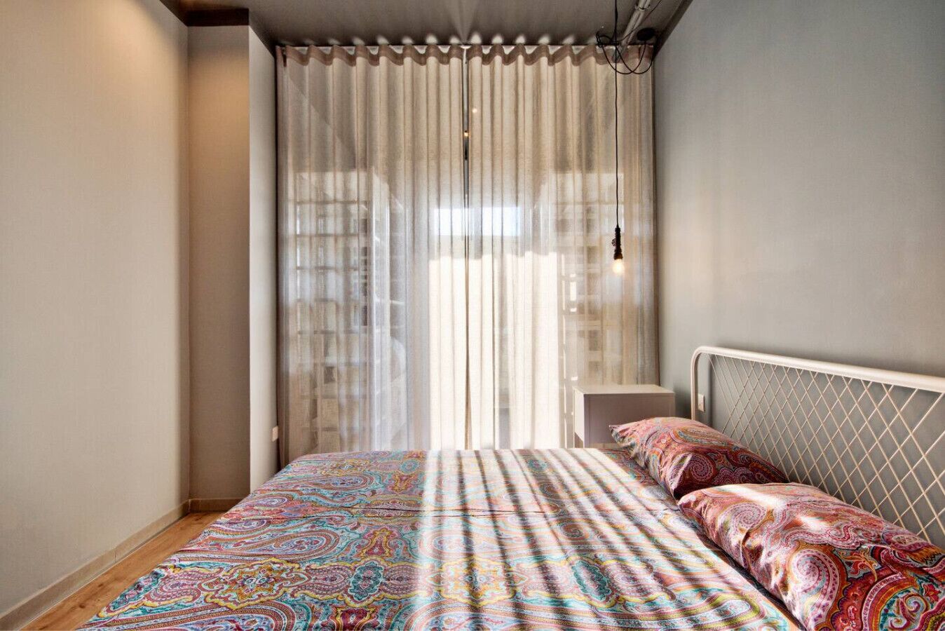Een slaapkamer met een bed, een nachtkastje en een raam met dunne gordijnen.