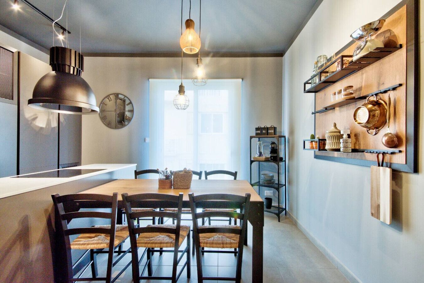A modern kitchen with a wooden dining table, chairs, pendant lights, and built-in shelves with kitchenware.