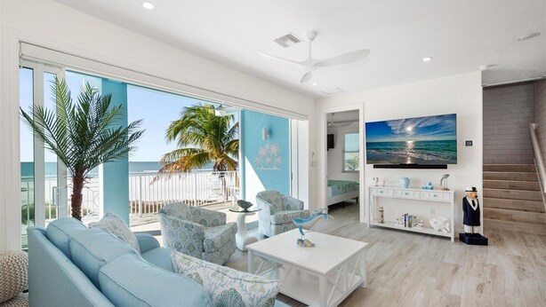 Een ruime woonkamer met een grote bank, een salontafel en een aan de muur bevestigde televisie. Er is een balkon met uitzicht op de oceaan en palmbomen.