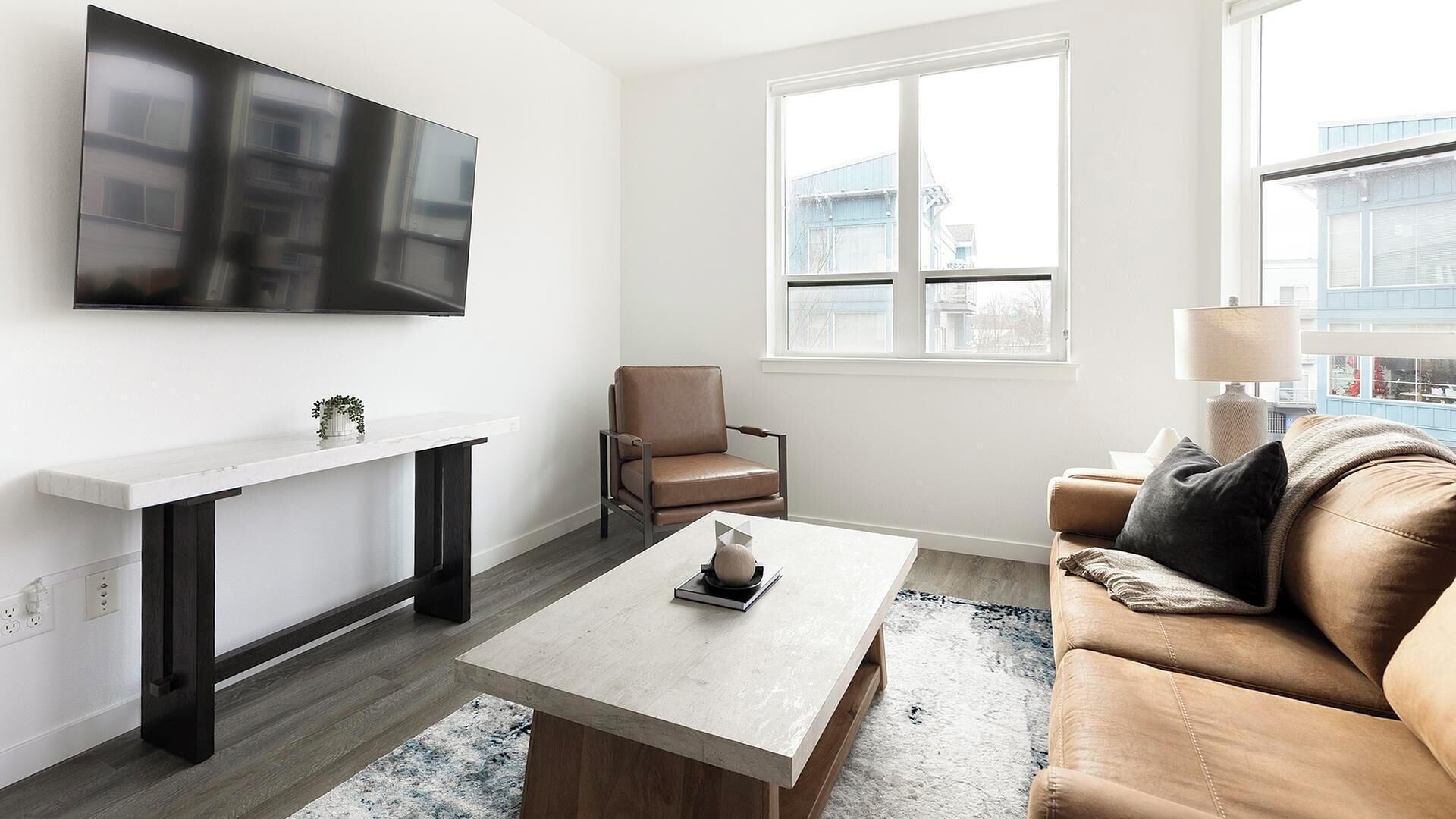 Ein modernes Wohnzimmer mit einem wandmontierten Flachbildfernseher, einer weißen Ablagetisch, einem braunen Ledersessel, einem hellen Sofa und einem Couchtisch mit einem dekorativen Gegenstand.