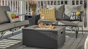 Eine Terrasse mit einer schwarzen gewobenen Couch, einer quadratischen Feuerstelle und einem weißen Geländer.