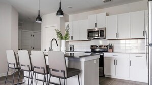 Een moderne keuken met witte kastjes, een roestvrijstalen koelkast, een magnetron en een koffiezetapparaat. Er staan drie stoelen met zwarte poten en witte kussens.