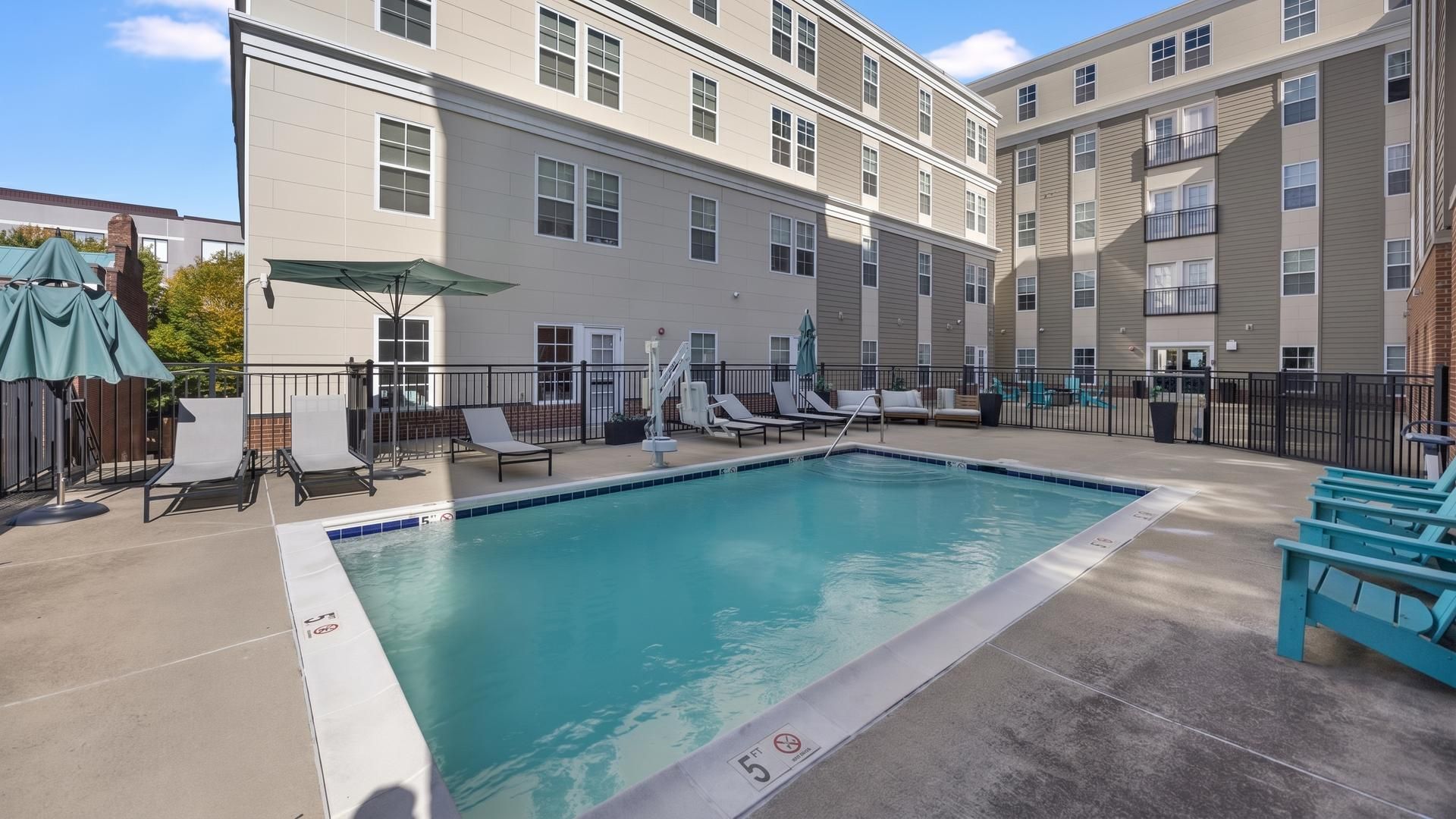 A swimming pool with lounge chairs and umbrellas, surrounded by a building with multiple windows and balconies.