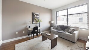 Ein modernes Wohnzimmer mit grauem Sofa, Couchtisch, Schreibtisch mit Stuhl und einem großen Fenster mit Jalousien.