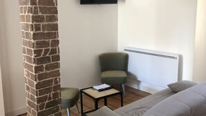 Un salon avec une colonne en briques, un canapé gris, un fauteuil vert, une petite table avec un livre et un téléviseur fixé au mur.