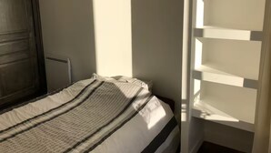 Un lit recouvert d’une couvre-lit à rayures, une porte en bois et un mur blanc sur lequel la lumière d’une fenêtre projette des ombres.