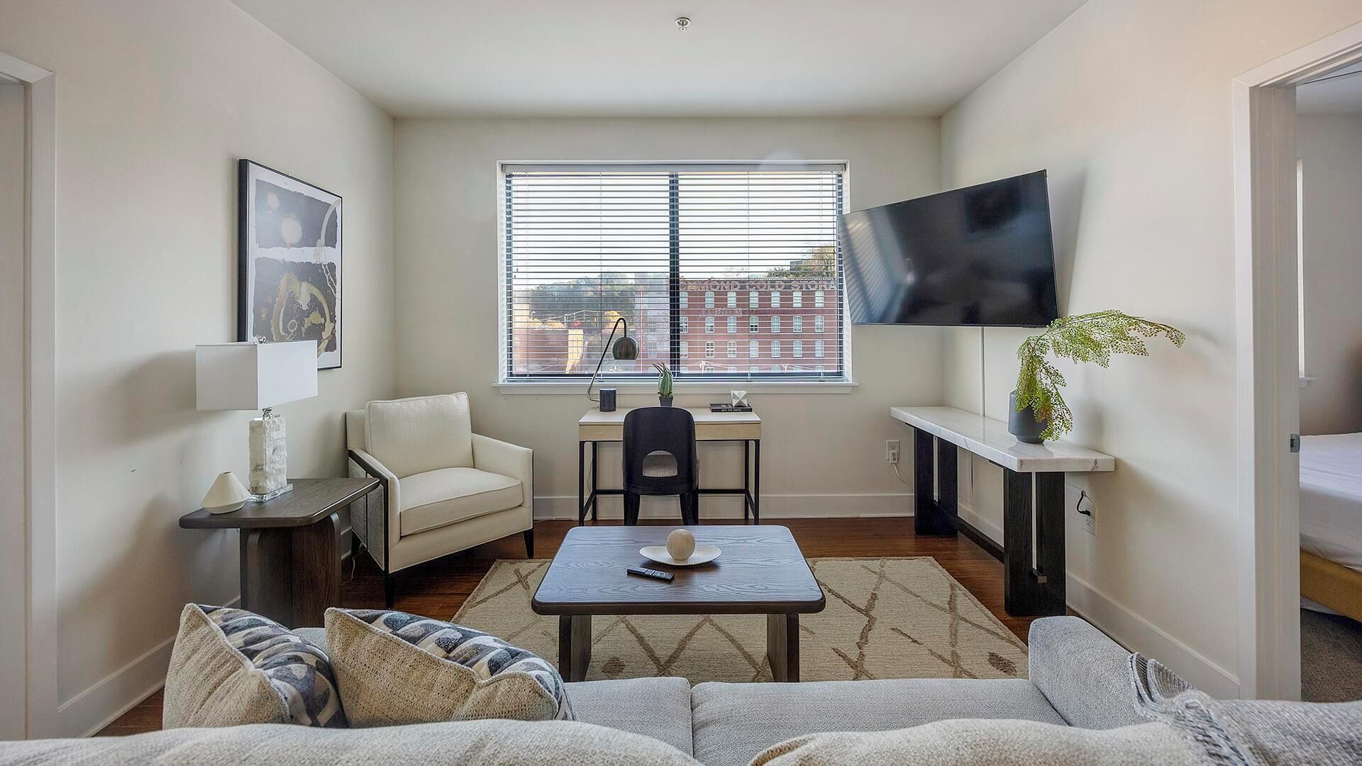 A compact living space with a sofa, a small table, a chair, a desk, and a TV mounted on the wall.