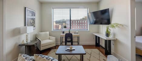 A compact living space with a sofa, a small table, a chair, a desk, and a TV mounted on the wall.