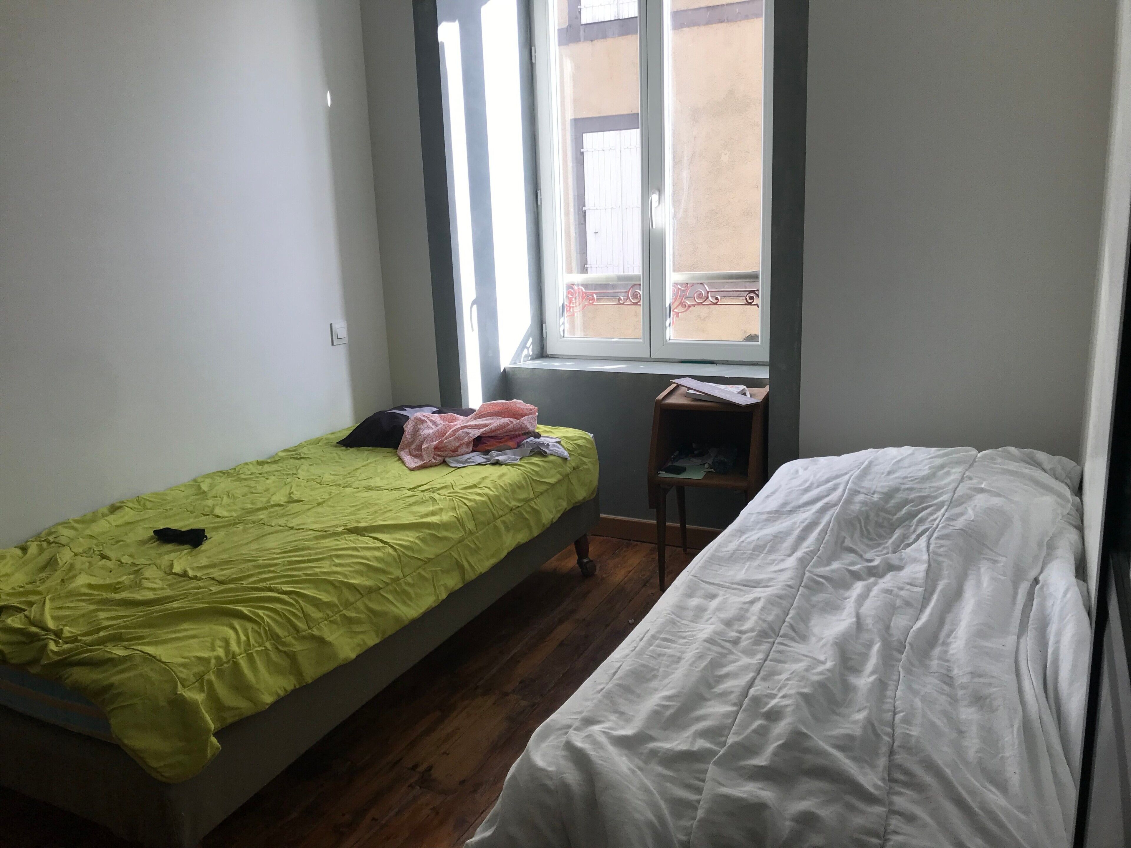 A small room with two single beds, a wooden floor, a window with a view, and a small wooden shelf.
