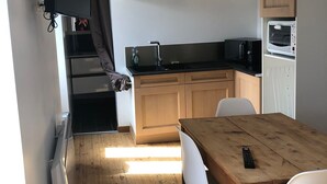 A compact kitchen with wooden cabinets, a built-in oven, a microwave, and a small refrigerator. There is a dining table with two chairs and a remote control on it. - Holiday Home 'T 2' with Wi-Fi (La Bourboule)