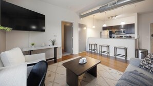 A modern living room with a flat-screen TV, a coffee table, and a sofa. There is a kitchen area with a microwave, oven, and bar stools. - Landing | Stunning 2BD, Rooftop Lounge, Courtyard (Richmond)