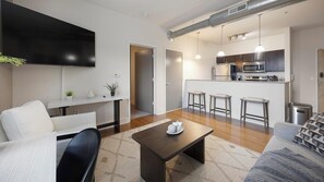 Ein modernes Wohnzimmer mit Flachbildfernseher, Couchtisch und Sofa. Es gibt eine Küchenzeile mit Mikrowelle, Backofen und Barhocker.