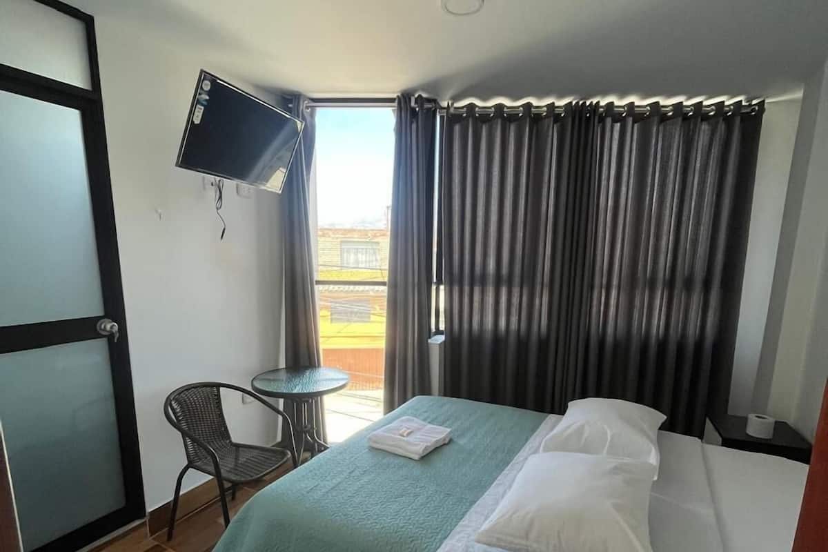 A hotel room with a bed, a chair, a TV mounted on the wall, and a window with curtains.