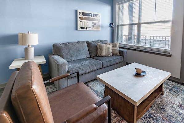 A modern living room with a grey sofa, a brown leather chair, a wooden coffee table, and a lamp on a side table. - Landing | Stunning 1BD, Courtyard, Gym (Enlow)