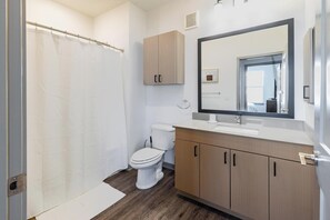A bathroom with a shower curtain, toilet, sink, mirror, and wooden cabinets. - Landing | Stunning 1BD, Courtyard, Gym (Enlow)