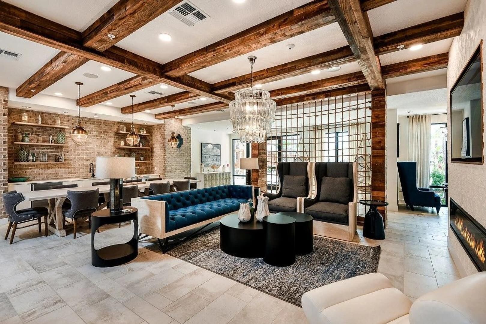 Ein modernes Wohnzimmer mit einem blauen Sofa, einem Couchtisch und einem Kamin. Es gibt eine Küchenzeile mit einer Ziegelwand und Holzbalken.