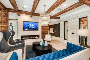 A modern living room with exposed wooden beams, a fireplace, a large TV, and a blue sofa. - Landing | Spectacular 2BD, Yoga Studio, Pool (St. Augustine)