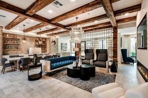 A modern living room with a blue sofa, a coffee table, and a fireplace. There is a kitchen area with a brick wall and wooden beams. - Landing | Spectacular 2BD, Yoga Studio, Pool (St. Augustine)