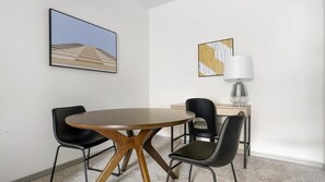 Une pièce moderne avec une table ronde en bois, quatre chaises noires, un bureau avec une lampe et deux tableaux abstraits accrochés aux murs.