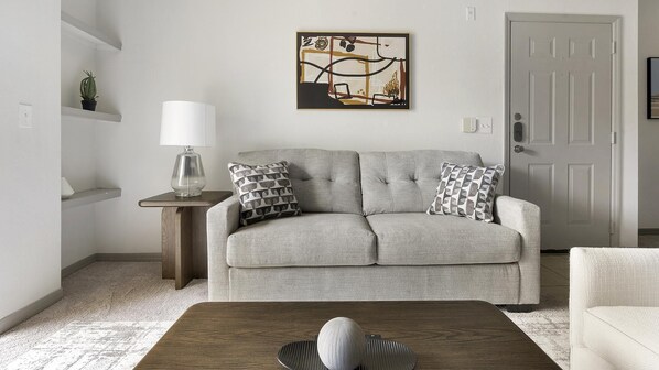 Un salon avec un canapé gris, deux coussins à motifs, une lampe au design épuré, un tableau accroché au mur et une table basse en bois surmontée d’une sphère décorative.