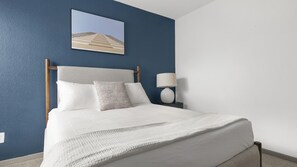 Un lit bien fait, avec du linge de lit blanc et une tête de lit grise, une lampe de chevet et un tableau encadré au mur.