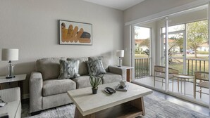 En moderne stue med en grå sofa, to sofaborde med lamper, et sofabord med en plante, og en stor skydedør med glas til en balkon.