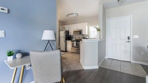 Et moderne køkken med rustfrie hårde hvidevarer, et spiseområde med en grå stol og et bord, samt en hvid dør.