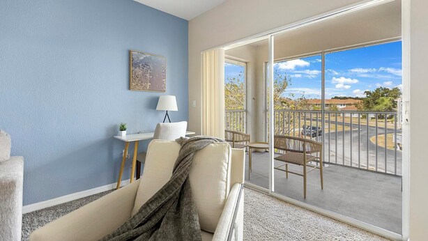 A modern living room with a sofa, a small table with a lamp, a framed picture on the wall, and a balcony with a view of the neighborhood. - Landing | Chic 2BD, Gym, Clubhouse (North Port)
