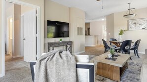 A modern living room with a flat-screen TV, a dining area with a table and chairs, and a kitchen in the background. - Landing | Chic 2BD, Gym, Clubhouse (North Port)
