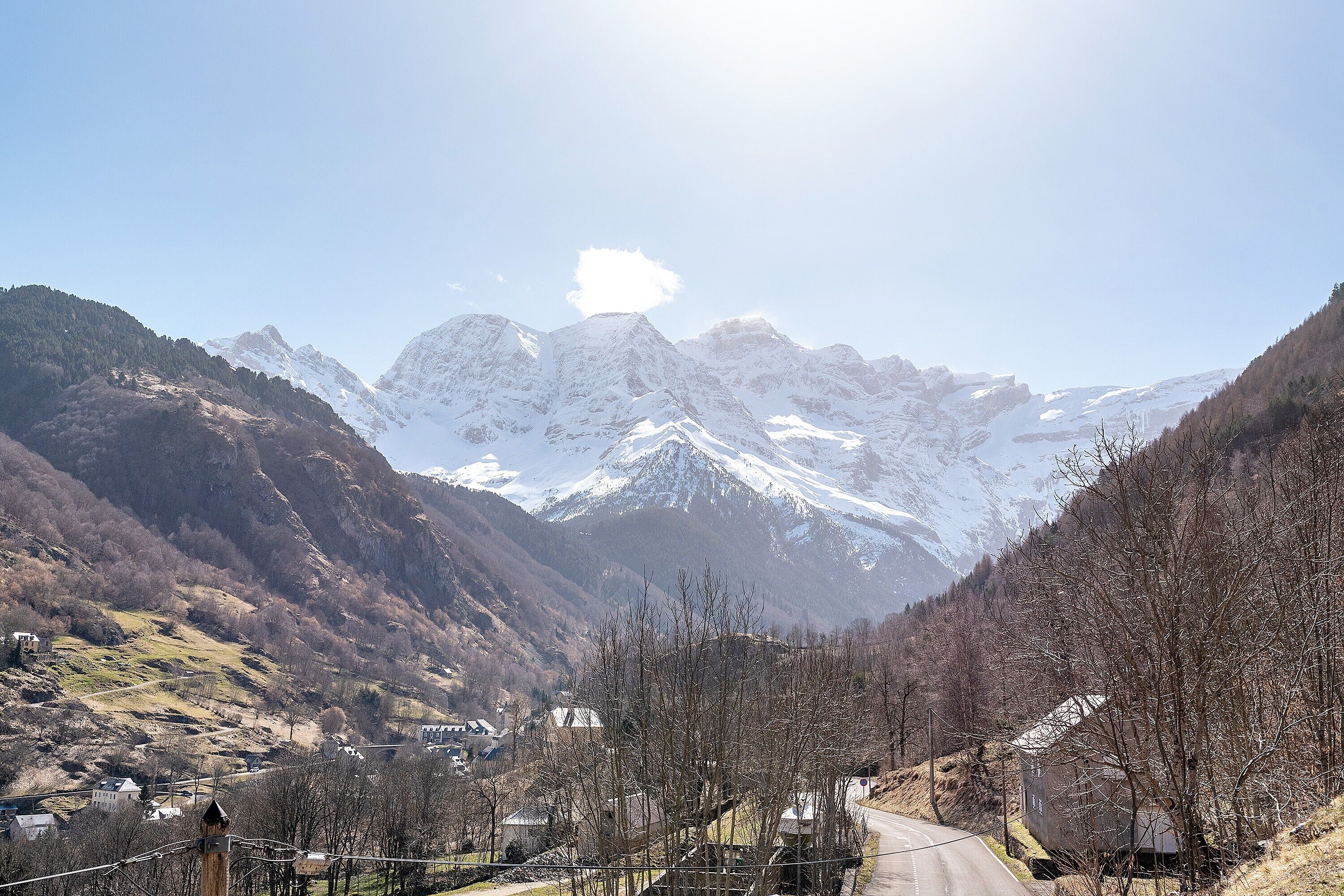 A mountainous landscape with snow-capped peaks, a clear sky, and a road leading through a valley with sparse vegetation and buildings.