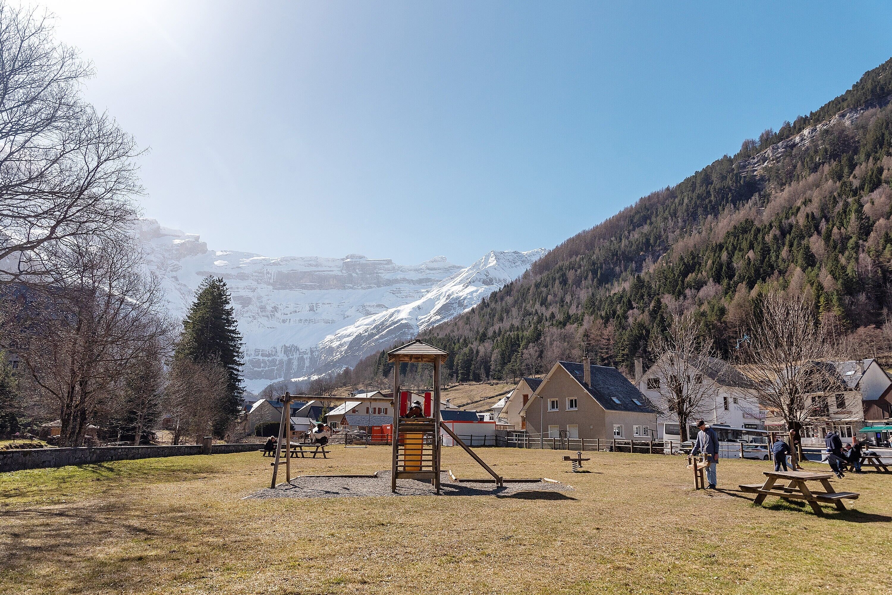 Ein Spielplatz mit Rutsche, Schaukeln und Klettergerüst in einem Park. Im Hintergrund sind Häuser und Berge zu sehen.