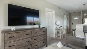 A modern living room with a large flat-screen TV, wooden dresser, and a small table with a lamp.