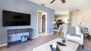 A living room with a TV, a coffee table, and a sofa. There is a kitchen in the background with a microwave and a sink. - Landing | Modern 1BD, Courtyard, Gym (North Port)