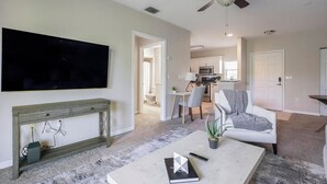 A living room with a flat-screen TV, a grey armchair, a coffee table with a book and a plant, and a dining area with a table and chairs. - Landing | Modern 1BD, Clubhouse, Gym (North Port)
