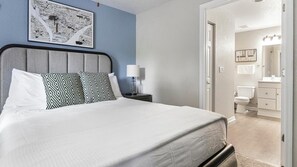 A neatly made bed with white bedding and patterned pillows, a bedside table with a lamp, and a framed map on the wall.