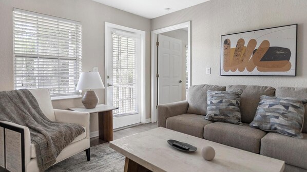 A living room with a sofa, armchair, and a coffee table. There is a door leading to another room and a window with blinds.