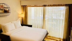 Une chambre d’hôtel comprenant un lit, une lampe de chevet, une fenêtre avec des rideaux et une étagère fixée au mur, ornée d’un cadre et d’objets décoratifs.