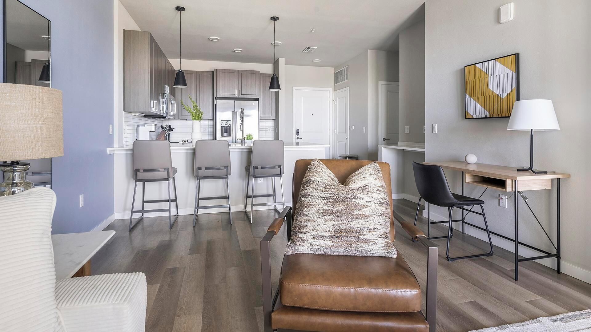 Una sala de estar moderna con un sofá de cuero marrón, una zona de comedor con una mesa y sillas de madera, y una cocina con armarios grises y electrodomésticos de acero inoxidable.